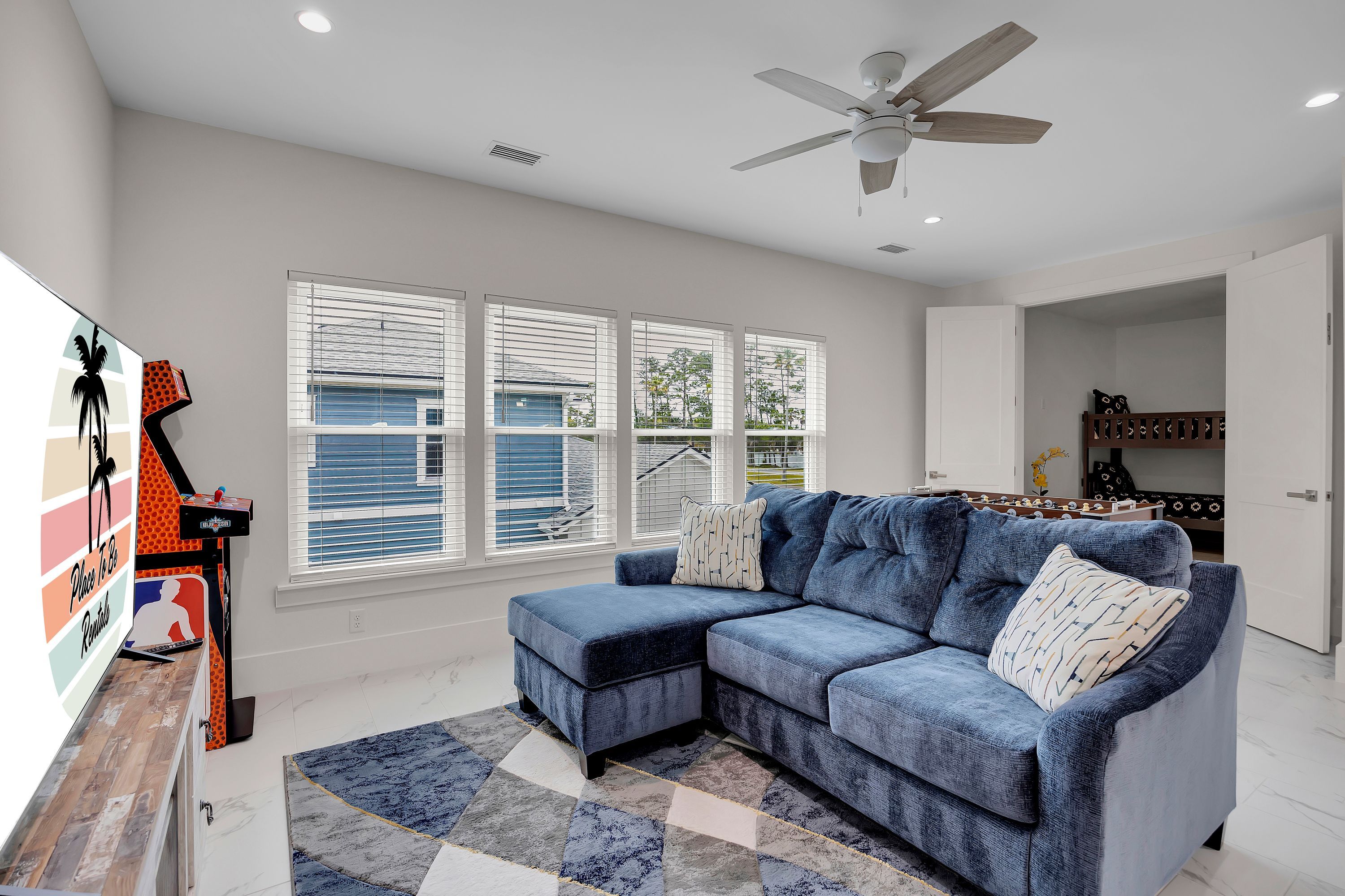 A modern living room with a sectional sofa, a ceiling fan, and large windows with blinds.