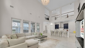 A spacious living area with a large sofa, a coffee table, and a dining area with chairs and a kitchen in the background. - Salt Air 6BR Game Room Walk to Beach Luxe (Ponte Vedra Beach)