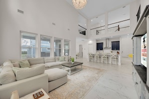 A spacious living area with a large sofa, a coffee table, and a dining area with chairs and a kitchen in the background. - Salt Air 6BR Game Room Walk to Beach Luxe (Ponte Vedra Beach)
