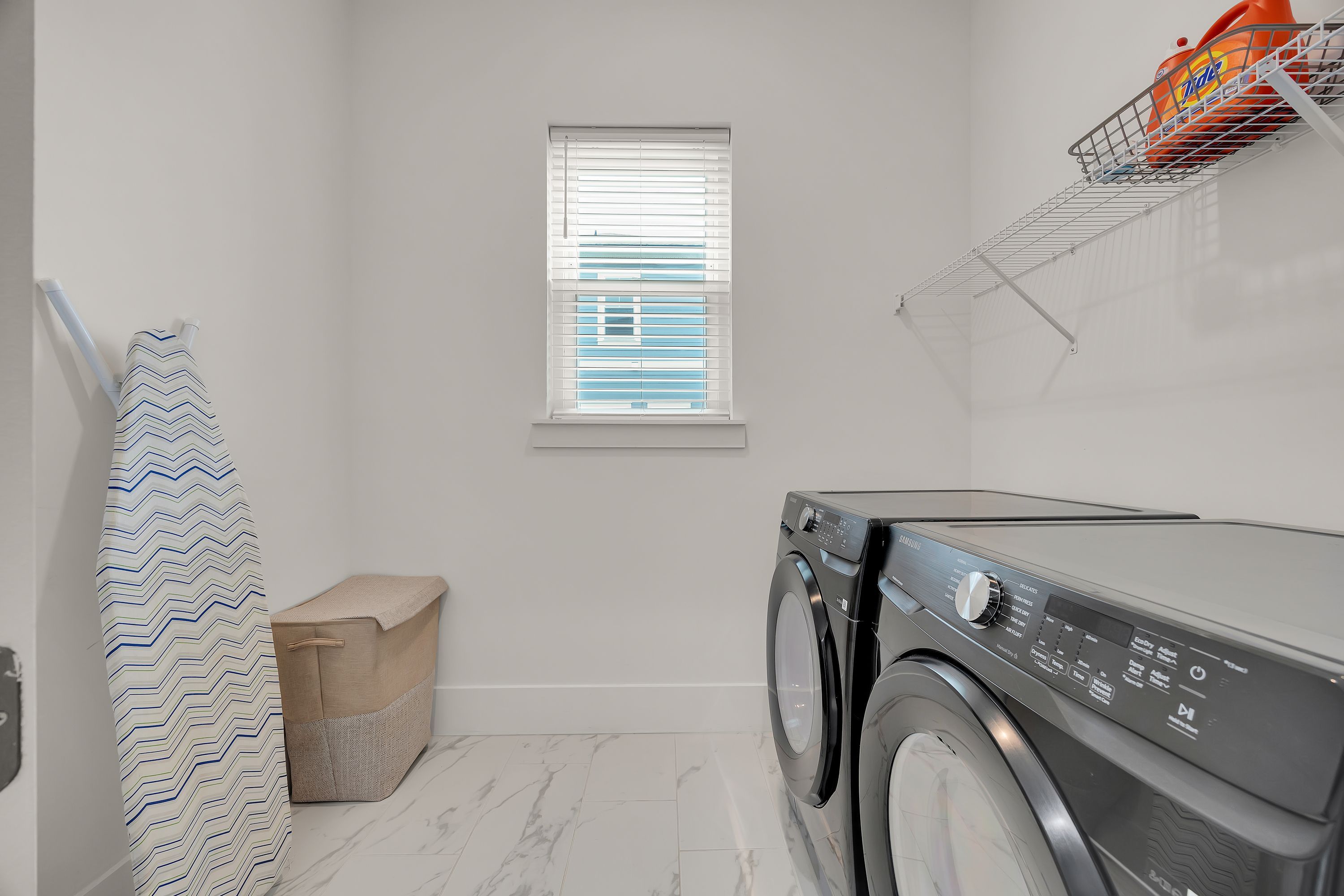 A laundry room with a washing machine, a window with blinds, a towel rack, and a storage bin.