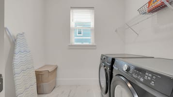 A laundry room with a washing machine, a window with blinds, a towel rack, and a storage bin.