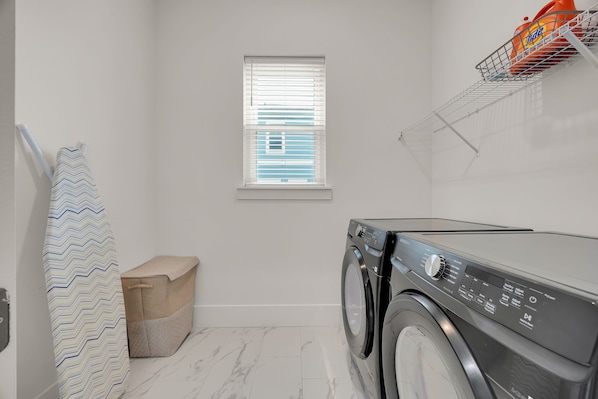 A laundry room with a washing machine, a window with blinds, a towel rack, and a storage bin.