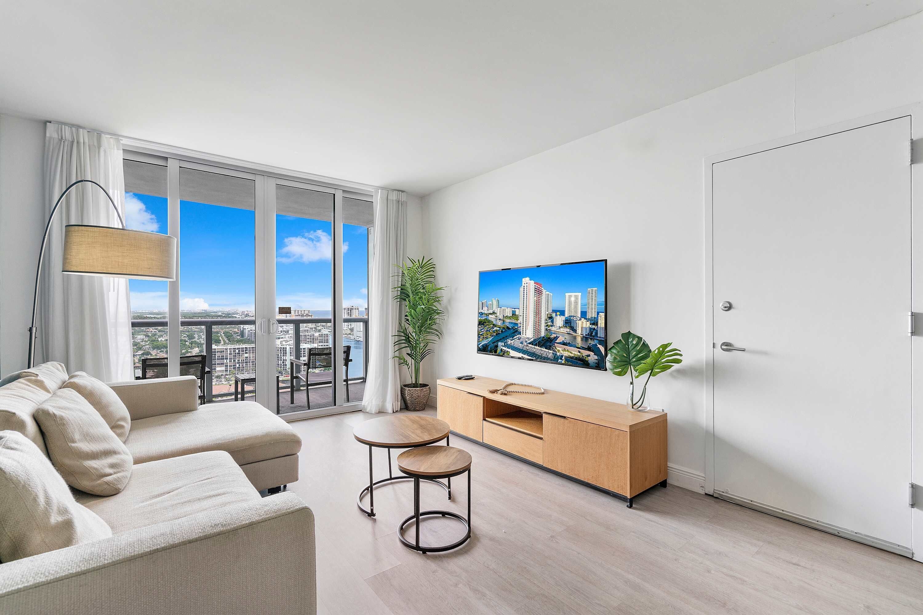 A modern living room with a sofa, a small table, a TV, and a cityscape view through large windows.