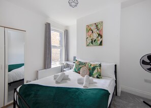 A hotel room with a bed, a desk, a chair, a window with curtains, a radiator, a wall-mounted artwork, and a standing fan. - Spacious 4BR House Near Elizabeth Line O2 Open (London)