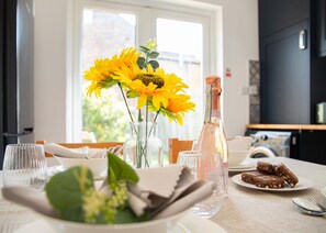 A dining table with a bottle of champagne, a plate of brownies, and a vase with sunflowers. - Spacious 4BR House Near Elizabeth Line O2 Open (London)