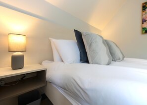 Una cama bien hecha con ropa blanca, una lámpara de noche y un cuadro colgado en la pared.