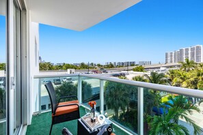 A balcony with a glass railing, a wicker chair, a small table with a bottle and glasses, and a view of a cityscape with palm trees and high-rise buildings. - Coastal Comfort 2BR Retreat Balcony Space (Sunny Isles Beach)