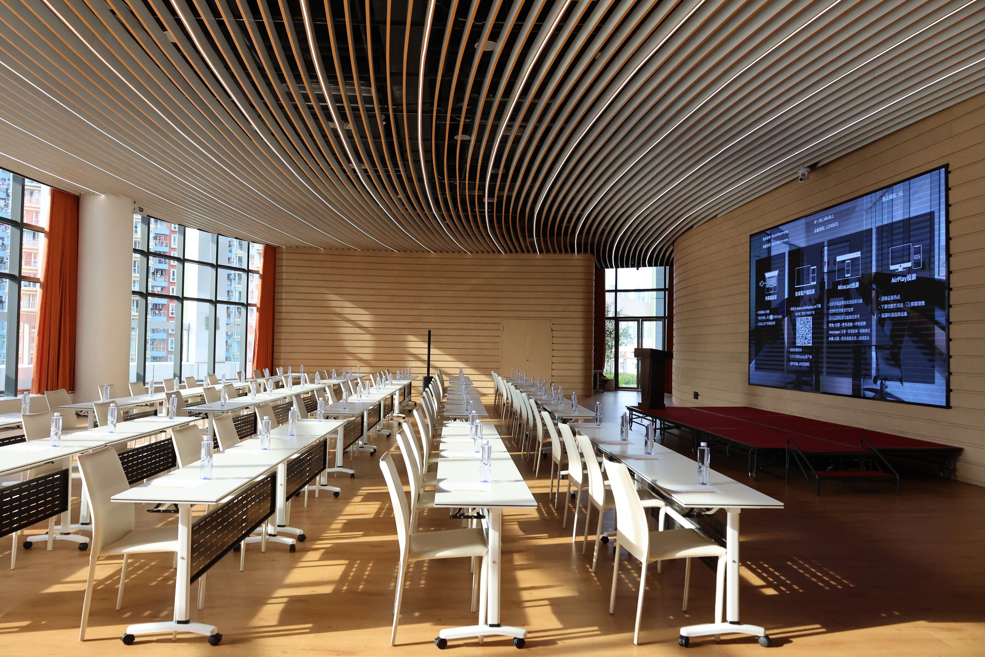 A conference room with rows of tables and chairs, a large projection screen on the wall, and large windows allowing natural light.