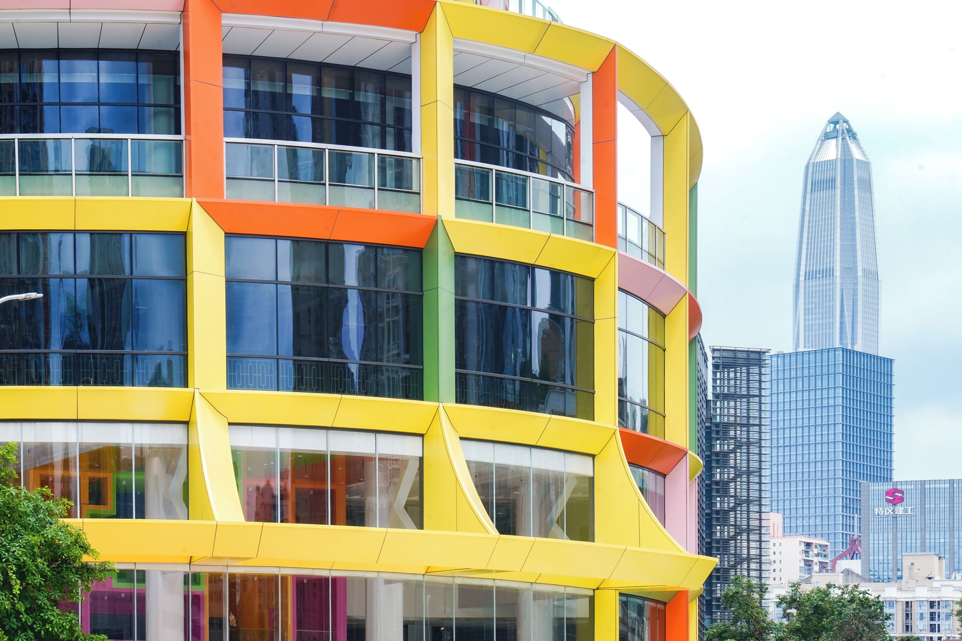 A modern building with a yellow and orange facade, large windows, and a reflection of a tall skyscraper in the background.