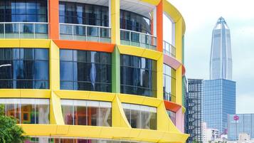 A modern building with a yellow and orange facade, large windows, and a reflection of a tall skyscraper in the background.
