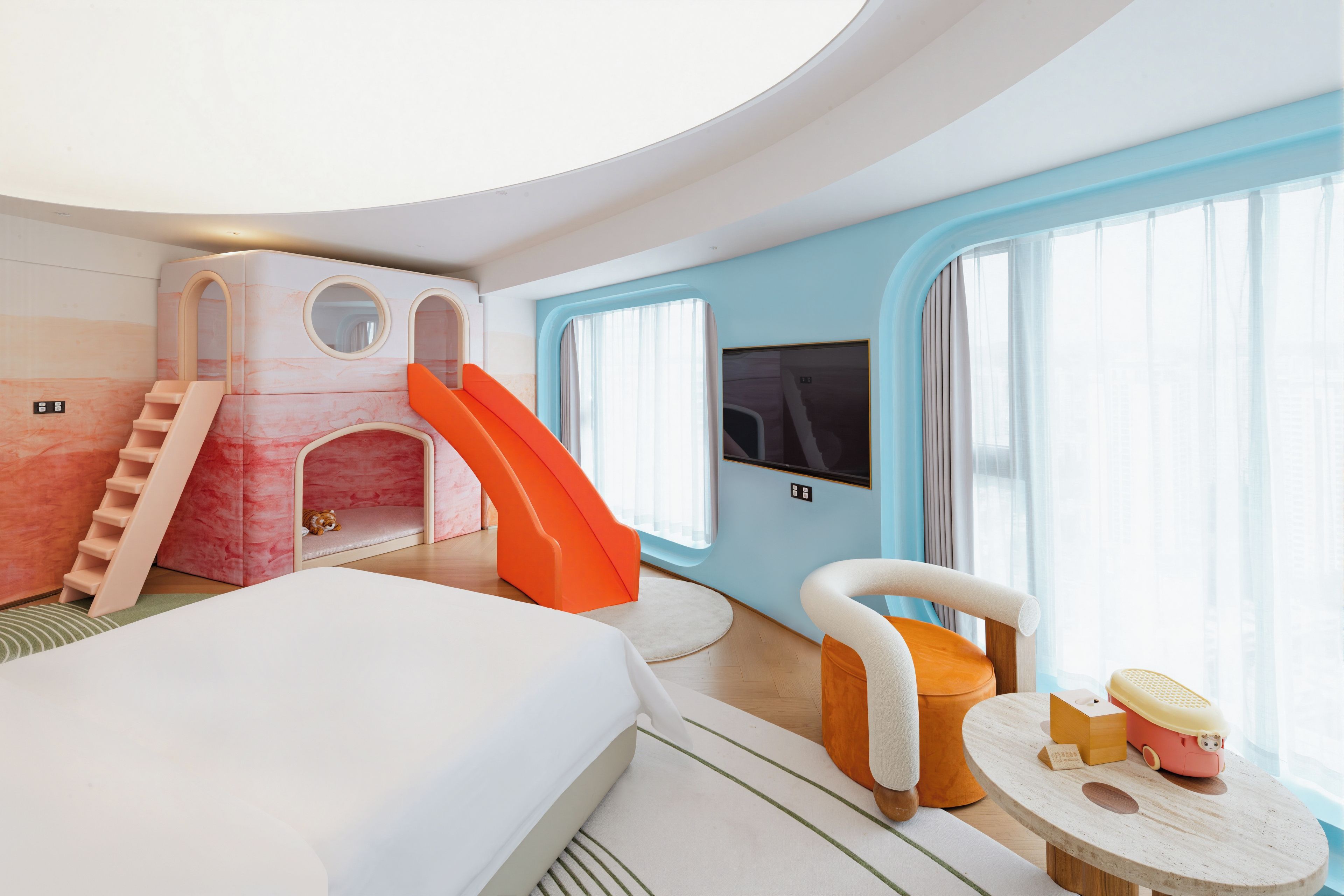 A modern hotel room with a bed, a play area with a slide, a TV, and a window with curtains.