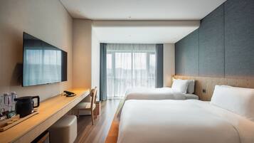 A hotel room with a flat-screen TV, a desk with a kettle, two beds, and a large window with curtains.