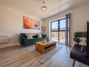 A modern living room with a green sofa, a wooden coffee table, and a large window with a view of buildings. - Derby City Centre Apt | Free Parking | Long Stays (Derby)