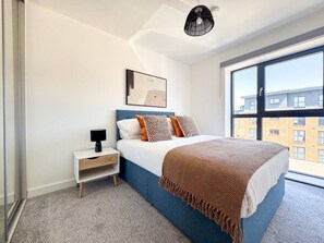 A modern bedroom with a large window, a bed with a patterned blanket, a bedside table with a lamp, and a wall-mounted artwork. - Derby City Centre Apt | Free Parking | Long Stays (Derby)