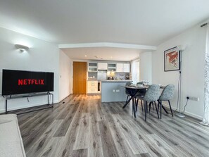 A modern living room with a flat-screen TV, a dining area with chairs, and a kitchen in the background. - Stylish 2BR Apt | Jewellery Quarter + Free Parking (Birmingham)