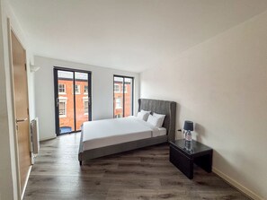 A bedroom with a bed, a nightstand, and a view of a red brick building through a window. - Stylish 2BR Apt | Jewellery Quarter + Free Parking (Birmingham)