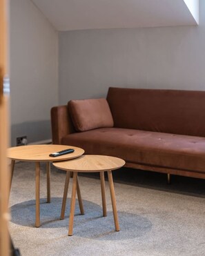 A room with a brown sofa, two round wooden tables, and a remote control on one of the tables. - Central 1 Bed | Prime Stratford Town Centre (Stratford-upon-Avon)