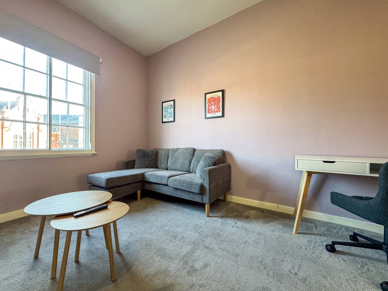 A room with a grey sofa, a small round table, a desk with a chair, and two framed pictures on the wall.