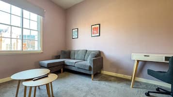 A room with a grey sofa, a small round table, a desk with a chair, and two framed pictures on the wall.