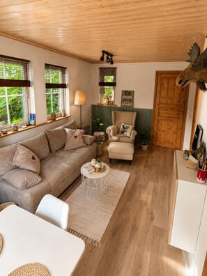 A cozy living room with a sofa, armchair, and a coffee table. There are windows with blinds, a lamp, and a door with a wooden frame.