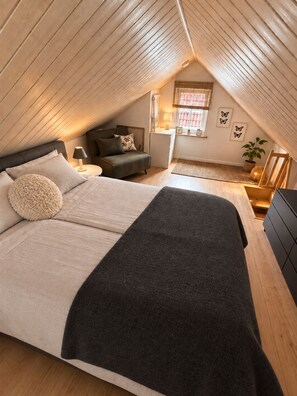 A bedroom with a sloped ceiling, a bed with a dark blanket, a nightstand, a small table, a window with blinds, and a potted plant.