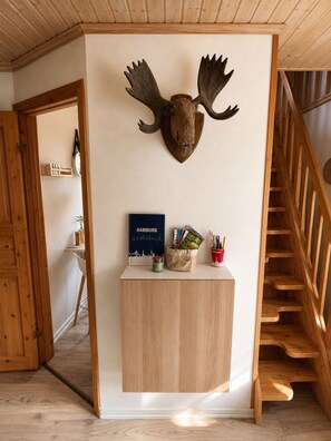 Ein Zimmer mit Holzbalkendecke, einem aufgehängten Elchgeweih an der Wand, einem Schreibtisch mit einem Hamburg-Reiseführer, einer kleinen Pflanze und einer roten Tasse mit Stiften.