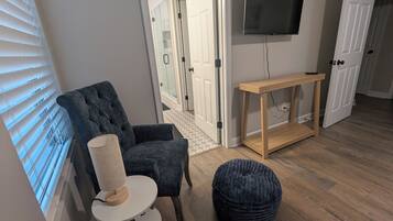 Ein Zimmer mit grauem Polsterruhesessel, einem kleinen weißen Tisch mit Lampe, einem runden Hocker, einem Holzkonsolentisch und einem an der Wand montierten Flachbildfernseher.
