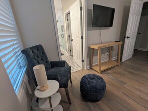 A room with a grey upholstered armchair, a small white table with a lamp, a round ottoman, a wooden console table, and a flat-screen TV mounted on the wall. - Private 3-bedroom house with Hot Tub near White Water Center and Belmont (Mount Holly)