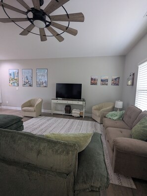 Ein Wohnzimmer mit Deckenventilator, einem Flachbildfernseher, zwei Sesseln, einem Sofa und einem Couchtisch.