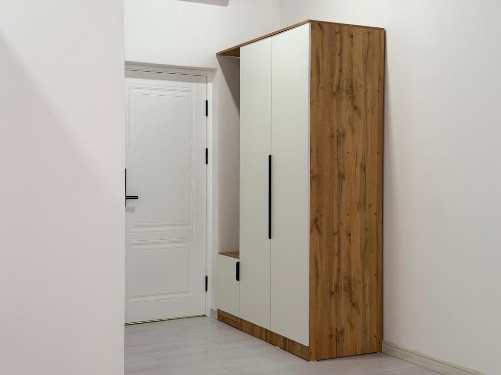 A wardrobe with a wooden finish and two doors, located in a hallway with a white door in the background.
