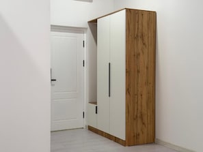 A wardrobe with a wooden finish and two doors, located in a hallway with a white door in the background.