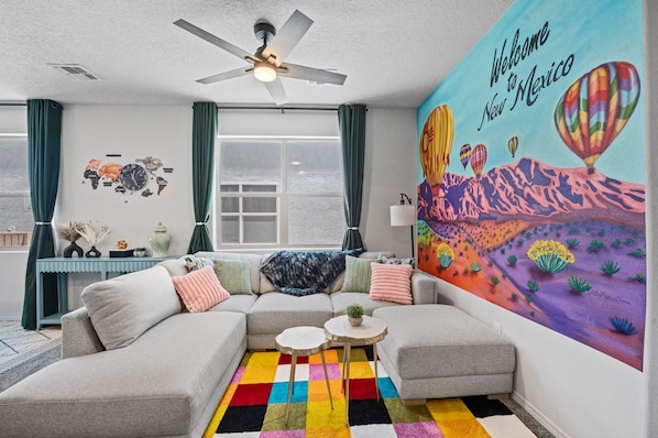 A modern living room with a sectional sofa, a colorful rug, a world map wall art, and a large mural depicting hot air balloons and cacti.
