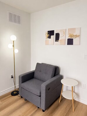 Een moderne woonkamer met een grijze bank, een vloerlamp, een klein wit bijzettafeltje en drie abstracte kunstwerken aan de muur.