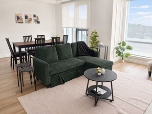 Ein modernes Wohnzimmer mit einer Eckcouch, einem Esstisch mit Stühlen, einem kleinen runden Tisch mit Fernbedienung und einem großen Fenster mit Blick.