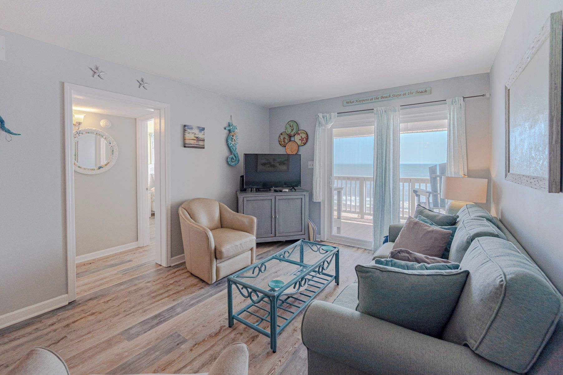 A living room with a sofa, armchair, and a TV on a cabinet. There is a balcony with a view of the sea.