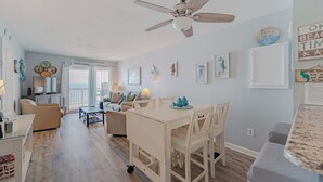 A spacious living area with a ceiling fan, a dining table with chairs, and a kitchen area in the background. - JL S-2C Sands Condos - Oceanfront - Community Pool (Kill Devil Hills)