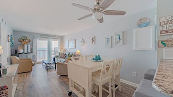 A spacious living area with a ceiling fan, a dining table with chairs, and a kitchen area in the background.