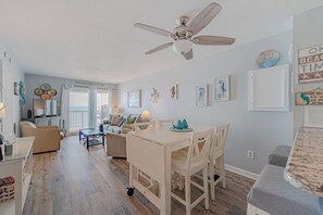 Een ruime woonkamer met een plafondventilator, een eettafel met stoelen en een keuken op de achtergrond.