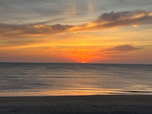 Een zonsondergang boven een rustige zee, met een strand op de voorgrond.