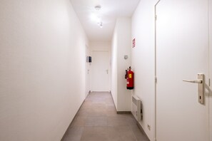 A narrow hallway with white walls and a tiled floor, leading to a door with a fire extinguisher mounted on the wall. - Apartment in Blankenberge With Sea Views (Blankenberge)