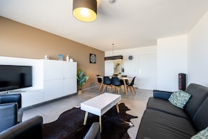 A modern living room with a flat-screen TV, a sofa, a coffee table, and a dining area with chairs and a table. - Apartment in Blankenberge With Sea Views (Blankenberge)