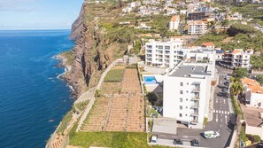 A coastal residential area with terraced vineyards, a modern building with a swimming pool, and a road with vehicles. - Azure Oasis Madeira by Atlantic Holiday (Câmara de Lobos)