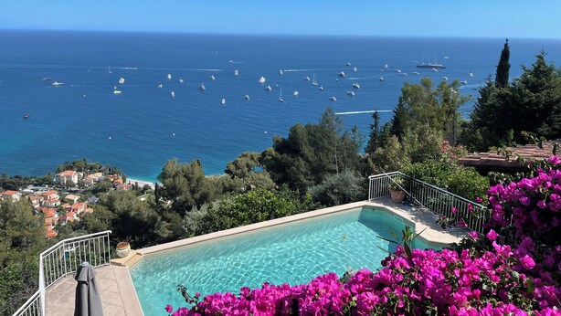 En pool med udsigt til havet og både, omgivet af grøn vegetation og blomster.