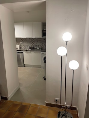 Ein Flur, der zu einer Küche mit weißen Schränken, einer Waschmaschine und einer Stehlampe mit drei Lampen führt.
