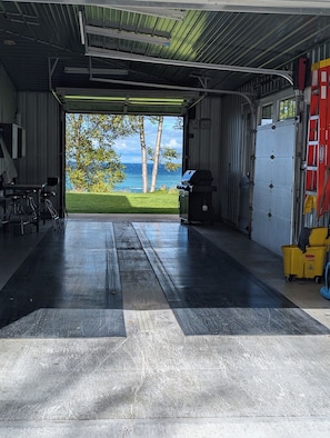A garage with a large open door leading to a grassy area with trees and a view of the ocean. Inside, there is a barbecue grill and a yellow trash bin. - The Ridge! Amazing view of Lake Superior, close to downtown & room for all (Grand Marais)