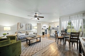 A modern living room with a sofa, coffee table, and chairs, a dining area with a table and chairs, and a kitchen in the background. - Elegant 2BR Apartment right by Cowboys Stadium! (Arlington)