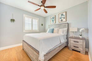 Ein Schlafzimmer mit einem hölzernen Bett, einem Nachttisch, einem Deckenventilator, einem Fenster mit Jalousien und zwei Wandpflanzen.