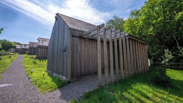 Een houten tuinhuis met schuin dak, omgeven door een grindpad en begroeiing.