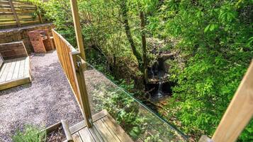 Une terrasse en bois dotée d’une rambarde en verre, qui offre une vue sur un espace naturel comprenant une petite cascade et une végétation luxuriante.