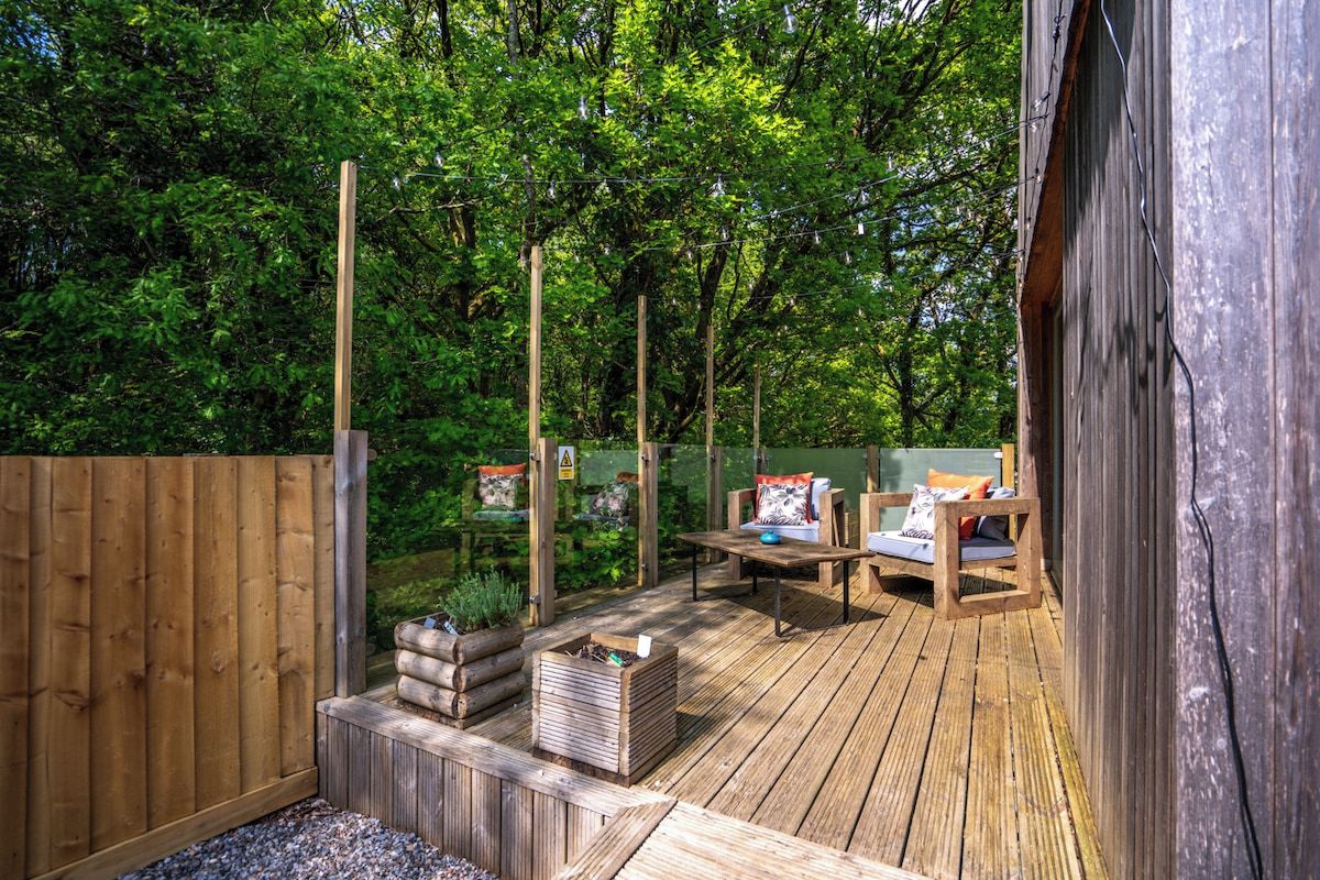 Terraza de madera con mobiliario exterior, rodeada por una valla de madera y abundante vegetación.